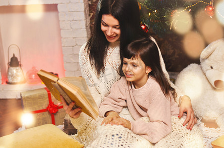 毯红摄影照片_美丽的年轻母亲和女儿正坐在圣诞树下, 在睡觉前阅读一本有童话故事的旧书.