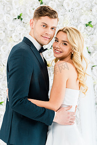 结婚领结婚纱摄影照片_微笑美丽的新娘拥抱英俊的新郎在白色花卉背景