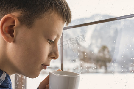 茶杯在手望着窗外没有在冬天雪山地森林景观的忧郁男孩