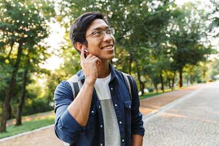 室外音乐摄影照片_微笑的亚洲男性学生在眼镜和耳机听音乐, 而看着远离