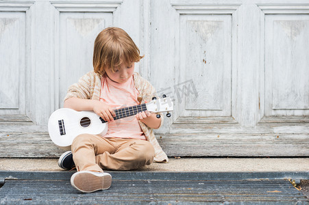 快乐的小男孩扮演他的吉他或夏威夷四弦琴，坐在户外的木门