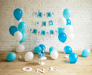 儿童节日礼物摄影照片_装饰一年生日与大量的蓝色和白色气球, 节日蛋糕和砖墙和地板上的铭文.