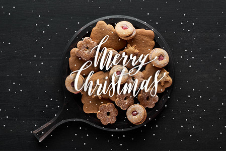 雪摄影照片_圣诞饼干放在黑色木制桌子上的平底锅上，上面印有圣诞快乐和降雪的图解