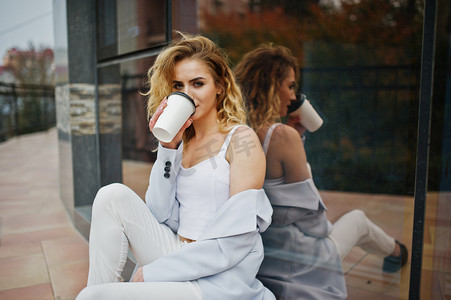 时尚的卷发金发模特女孩穿着白色与杯咖啡