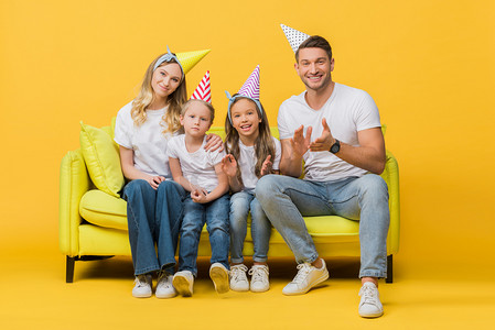 圣诞快乐应为摄影照片_戴着生日派对帽的快乐家庭在黄色的沙发上欢呼