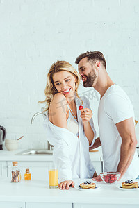 创意橙汁海报摄影照片_微笑的男人拥抱快乐的女朋友拿着草莓在厨房