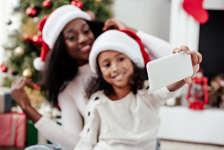 新科技智能摄影照片_微笑的非洲裔美国人母亲和女儿在圣诞老人帽子自拍在智能手机在家里