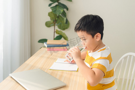 亚洲小男孩在使用笔记本电脑、坐在办公桌前做作业时喝水的肖像。电子学习和教育概念.