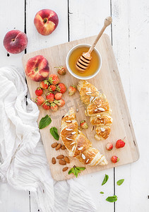 wood摄影照片_Freshly baked almond croissants with garden strawberries, peaches, mint and honey on serving board over white rustic wood backdrop