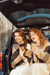 有选择的焦点笑妇女包裹在毯子里, 拿着一次性咖啡杯, 坐在汽车后备箱在街上