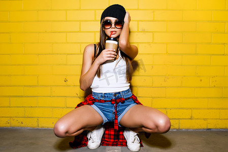 黄色格子背景摄影照片_美丽的年轻性感的女孩喝咖啡，坐在附近的黄色墙壁背景在太阳镜，红色格子衬衫，短裤.
