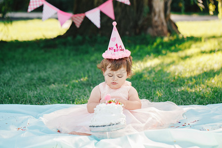 她笑的好开心摄影照片_深棕色的眼睛，穿着粉红色的短裙，与美食蛋糕外公园，蛋糕砸在她第一次生日可爱可爱高加索女婴的肖像