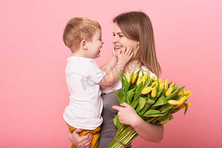 年轻的母亲抱着坐在地板上的小儿子, 背景是粉红色的。妈妈拿着一束春天的黄色花朵。关怀和人际关系以及家庭观念。母亲节