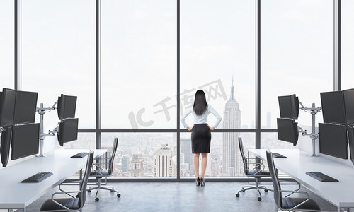 out摄影照片_Rear view of a lady in formal suit who is looking out the window in the modern panoramic office with New York view. White tables equipped with modern trader's stations and black chairs.