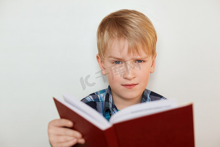 人脸表情和情绪。儿童和教育。有吸引力的小男孩，用公平的头发看书已经厌倦了作业有愤怒的表情的特写镜头。无聊的学校研究