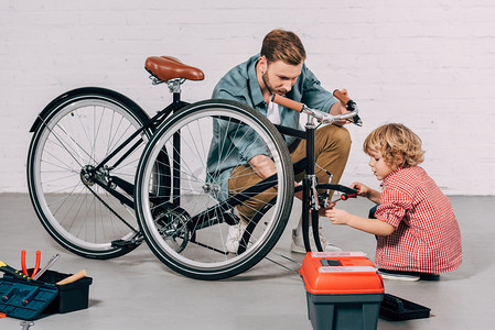 放心商业摄影照片_在车间里, 帮助小男孩在工具箱里的工具箱附近修理自行车的男子