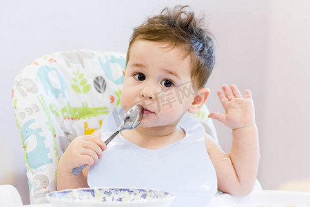 嘴馋的孩子摄影照片_微笑的孩子在厨房吃的食物。1 年岁宝宝吃他们自己的汤。第一次的固体食物，如何教孩子用勺子吃饭。可爱的宝宝，吃午饭用勺子吃饭