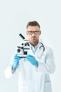 英俊的严肃的科学家在眼镜举行显微镜, 看着相机孤立的白色