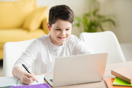 开朗的男孩使用笔记本电脑和写在抄本，而在家里做功课