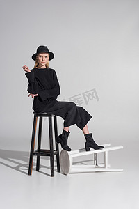 美丽的高加索金发碧眼的模型在时尚的黑色衣服和帽子坐在椅子上白色