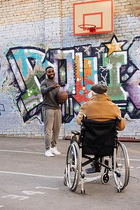 坐在轮椅上的高级残疾人的后视镜看着快乐的非洲裔美国人在街上打篮球