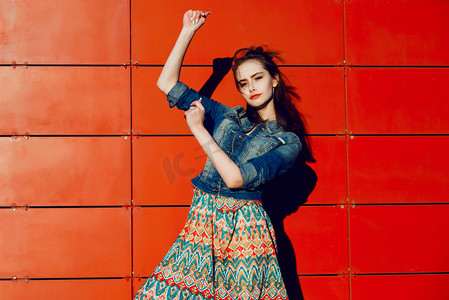 Young teenager girl having fun, posing and smiling near red wall background in skirt and jeans jacket on the sunset.