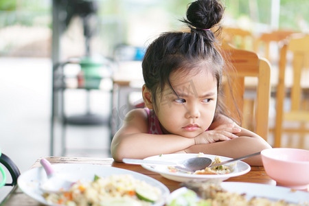 生闷气摄影照片_亚洲小孩可爱或小孩厌食症或伤心无聊的食物，或困倦的空余食物，撑着身子或面颊靠在木桌上吃早餐或午餐