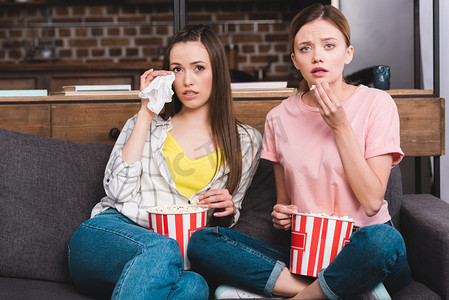 两个心烦的女性朋友与桶爆米花看电影在家里的沙发上 
