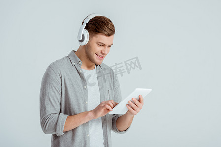 英俊的男人在耳机听音乐, 并使用数字平板电脑孤立在灰色