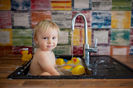 浴室儿童摄影照片_可爱的微笑的婴儿洗澡在厨房水槽。孩子们在阳光明媚的厨房里玩泡沫和肥皂泡, 厨房里有橡胶鸭和玩具。小男孩洗澡, 用水有趣