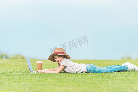 可爱的孩子在帽子微笑, 而在绿色草地上使用笔记本电脑