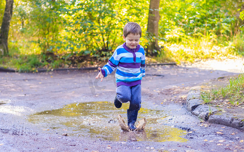 一个小男孩在水坑里跳。一个穿橡胶靴的男孩快乐的童年。雨后水坑。温暖的夏日夜晚.