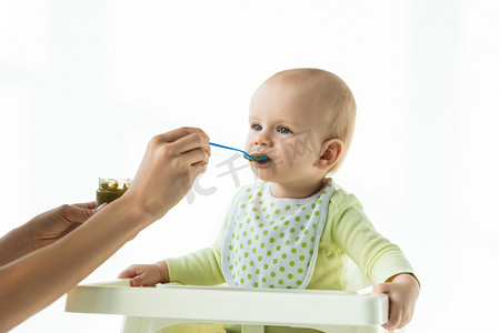 vegetable摄影照片_Mother with jar of vegetable baby nutrition and spoon feeding infant on feeding chair isolated on white