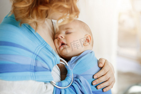 关闭了画像的无辜新生男婴母亲在婴儿背带的胸部上有甜美的梦。妈妈看着她的孩子用爱和温柔.