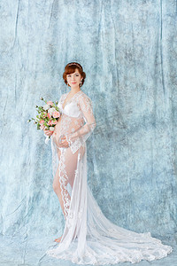 手捧鲜花，站在闺房服饰 （睡衣） 在蓝色背景上的孕妇.