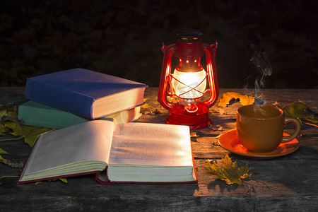 春天黄昏摄影照片_书的翻开，灯和一杯热咖啡放在黑暗的森林的旧木桌上。落黄枫叶。复古和古老的概念。秋天的心情.