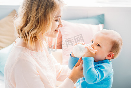 婴儿与奶瓶摄影照片_快乐的母亲在家喂养她的孩子与婴儿奶瓶