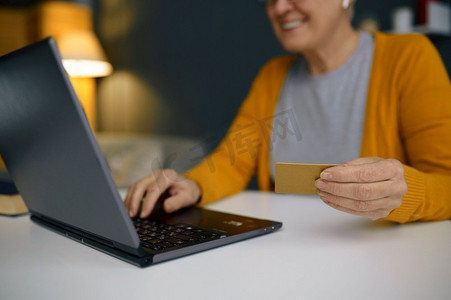 一位头发花白的成熟女性在家里用笔记本电脑使用银行信用卡进行在线支付。成熟女性在笔记本电脑上进行在线支付