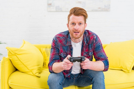 肖像兴奋的年轻人坐在黄色沙发玩电子游戏