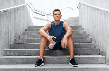 健身，运动和人的概念—疲惫的年轻人与蛋白质摇瓶坐在楼梯。运动员坐在楼梯上瓶子里