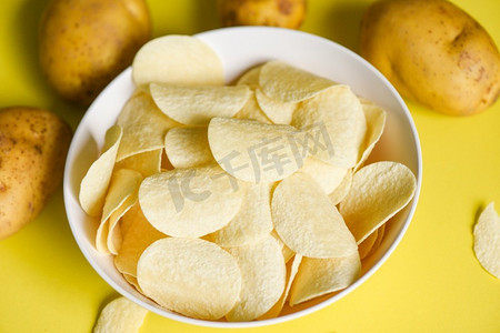 厨房桌子上放着酥脆的薯片和黄色背景的新鲜生土豆，碗里放着薯片零食