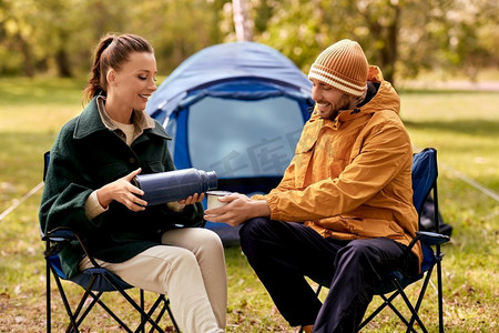 露营、旅游和旅行概念—愉快的夫妇与热水瓶倒茶杯在帐篷营地。快乐的夫妇与热水瓶倒茶在帐篷营地