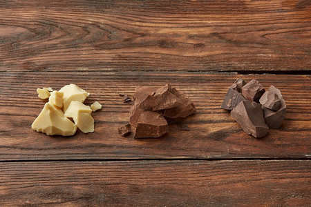 从上面开始，一组白巧克力、牛奶巧克力和黑巧克力被碾碎成块，排成一排放在木桌上。木质表面上的各式巧克力