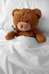医学，保健和儿童概念—生病的泰迪熊玩具头与温度计躺在床上。生病的泰迪熊玩具头与温度计在床上