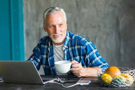 微笑的男人拿着咖啡杯，拿着笔记本电脑桌子望向别处