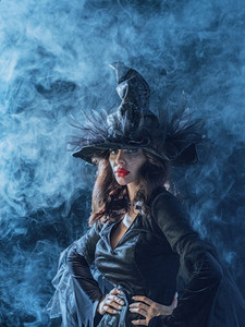 身穿万圣节女巫服装的美女在迷雾中戴着帽子，蓝色神秘的光芒。穿着万圣节女巫服装的女人