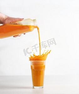 飞溅水花摄影照片_妇女们用手将自制的黄色果汁从瓶子里倒入玻璃中，白色背景上有飞溅的液体。准备了健康的奶昔饮料。前视图。