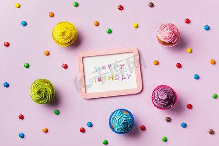 生日快乐背景粉色摄影照片_生日快乐石板周围有五颜六色的宝石松饼粉色背景