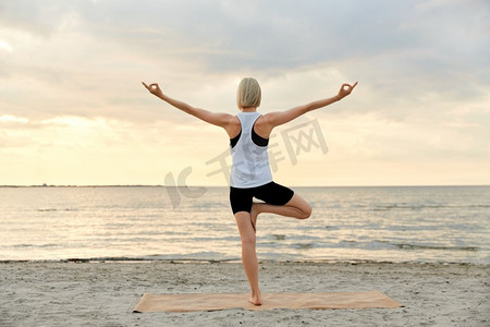 户外运动老外摄影照片_健身，运动，和健康的生活方式概念—妇女做瑜伽树姿势在海滩超过日落。妇女做瑜伽树姿势在海滩在日落