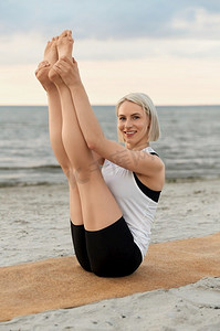 户外运动老外摄影照片_健身、运动和健康的生活方式概念—妇女做瑜伽船姿势在海滩超过日落。妇女做瑜伽船姿势在海滩